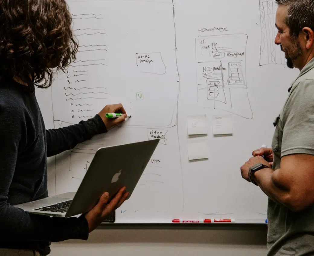 Woman explaining something to a man next to a whiteboard where she is writing with a black marker