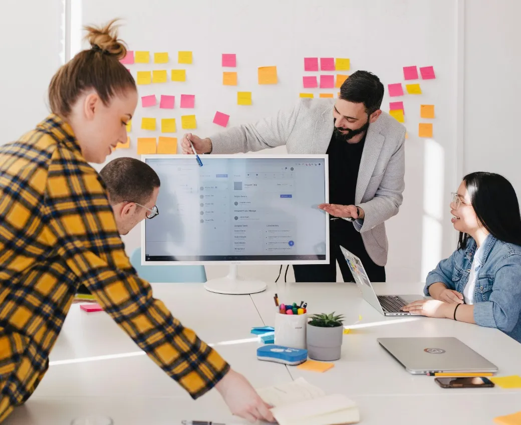 Man presenting on monitor to coworkers, with colorful stickers on whiteboard, creating a vibrant atmosphere at work.