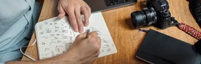 Man sitting at a table drawing sketches in a notebook, with a camera and a laptop beside him.