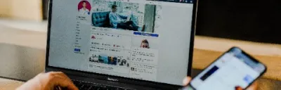 Social media profile being viewed on a laptop, with the observer holding a phone in their right hand