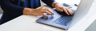 Woman in a blue blouse sitting at a table typing on a laptop.