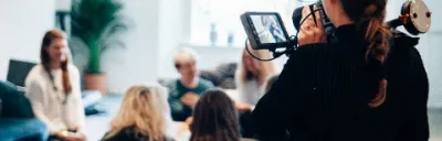 Woman holding a camera, recording people sitting in armchairs having a conversation.